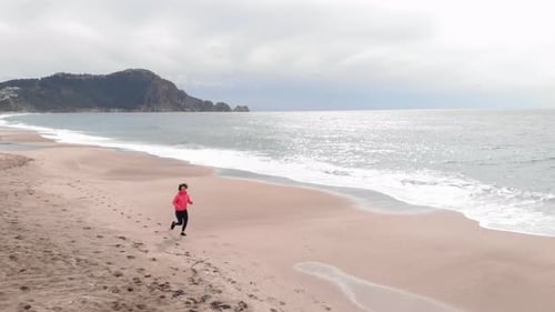 Young female athlete in orange jacket is running along seashore on sandy beach at stormy weather