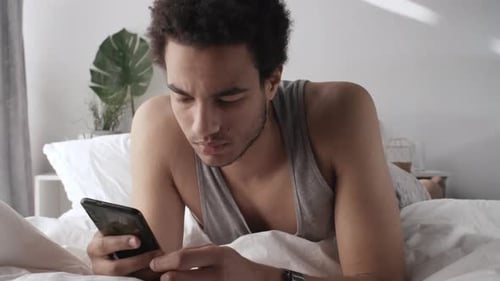 Man Lies in Bed Checking Phone and Smartwatch