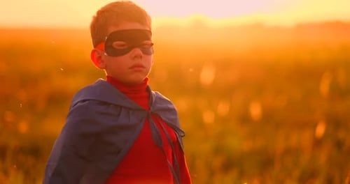 A Child in the Costume of a Superhero in a Red Cloak Runs Across the Green Lawn Against the Backdrop