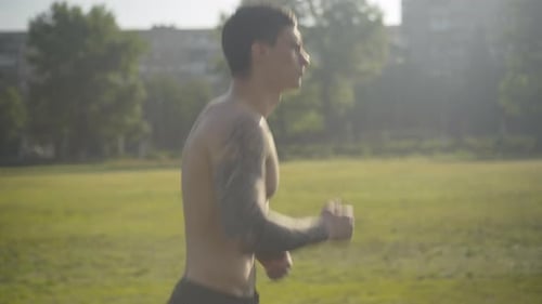 Middle Shot Side View of Sportive Caucasian Man with Tattooed Hand Jogging in Sunrays Outdoors