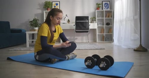 Woman Looks at Phone on Exercise Mat Indoors