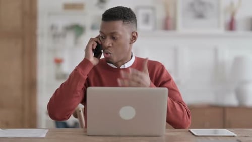 Angry African Man Talking on Phone while using Laptop in Office