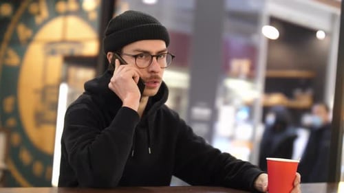 A Man in Black Clothes and a Hat is Talking on the Phone in a Shopping Mall
