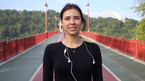 Portrait of a Sports Woman on the Bridge. Beautiful Brunette in Black Dress with Earphones Smiles