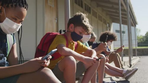 Grupo de niños que usan mascarillas y usan teléfonos inteligentes mientras están sentados juntos
