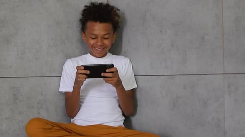 Front View of Cheerful Boy is Using Smartphone While Sitting in Light Interior Spbi