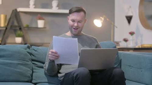 Man Sitting on Couch Reading Paper and Using Laptop