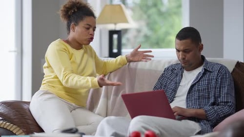 Esposa afro-americana irritada discutindo com marido absorvido trocando mensagens on-line no laptop