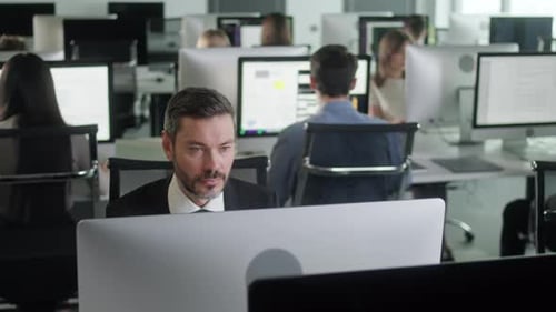 Serious Young Bearded Man Working on Decktop Computer While Working in Big Open Space Office