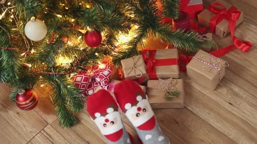 Feet in Funny Christmas Socks Santa Face Wooden Floor Under Fir Tree Gift Boxes