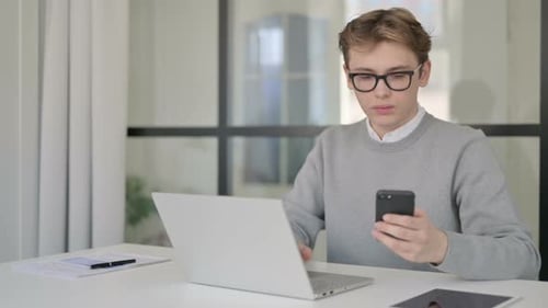Young Man Using Smartphone While Using Laptop in Modern Office