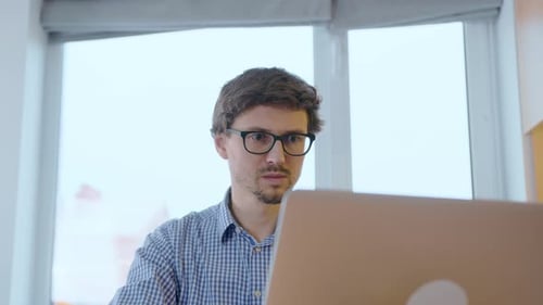 Man in glasses works at a laptop against the background of a window. Freelancer working remotely.