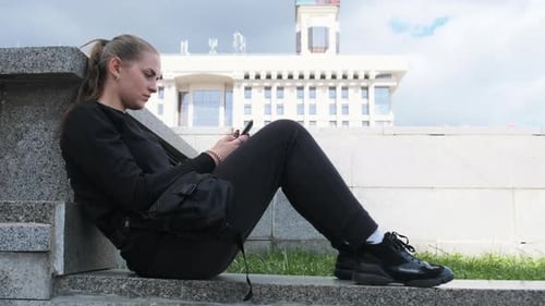 Young Woman Uses Smartphone While Sitting on the Parapet in the Street Park