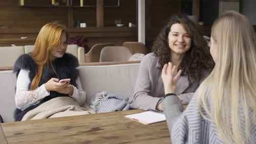 Three Caucasian Girlfriends Sitting at the Table in the Cafe. One Girl Holding Phone, Two Another