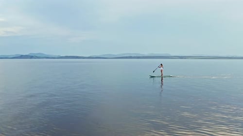 Drone Shot of a Young Woman with Long Hair Floating on a Paddleboard on a Mountain Lake at Sunset