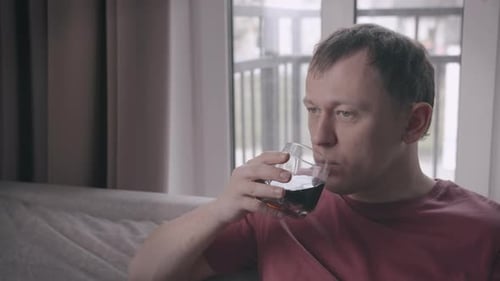 Young Man Sitting on Sofa with Glass of Drink in Hand Window Background