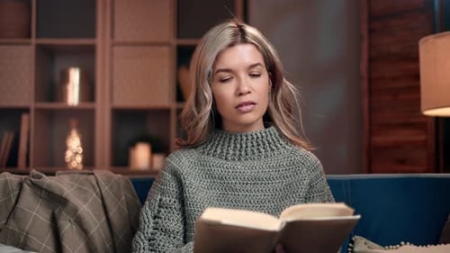 Glückliche blonde Frau in warmem Pullover liest Papierbuch entspannend am Abend zu Hause Gemütliches Interieur