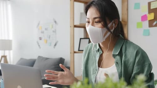Asian woman wearing face mask using laptop talk to colleagues in video call working from home.