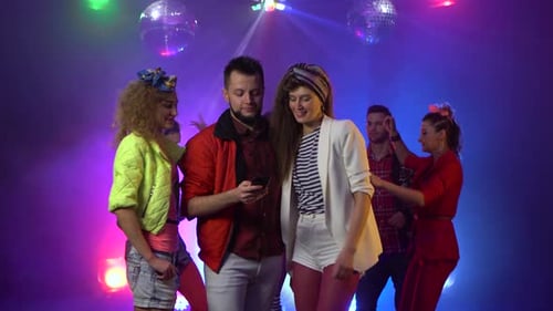 Friends in the Club Are Considering Something on the Phone Around People Are Dancing in the Smoke