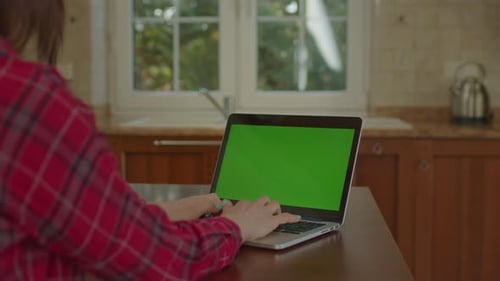 Over the Shoulder View of Woman Working on Laptop with Green Screen Sitting in the Kitchen