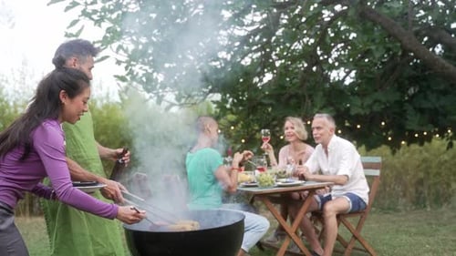 Mature Friends Enjoying Outdoor Summer Barbeque In Garden Cooking Grilled Corn and Having Fun