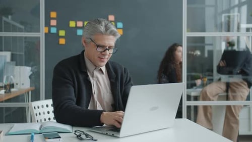 Empresario exitoso que usa una computadora portátil escribiendo y sonriendo en la oficina mientras las mujeres hablan de fondo