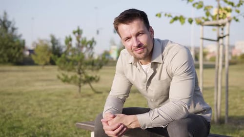A Young Caucasian Man Smiles at the Camera As He Sits in a Park in an Urban Area on a Sunny Day