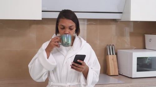 Woman in Robe Drinks Coffee and Uses Smartphone