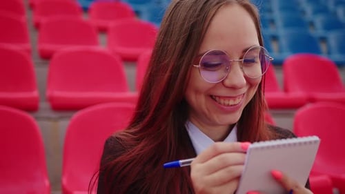 Young Woman in Glasses with Notepad Pen Sitting on Stadium Bleachers Alone