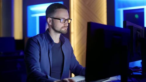 A Man with Glasses Sits and Thinks Typing on a Computer in the Office at Night