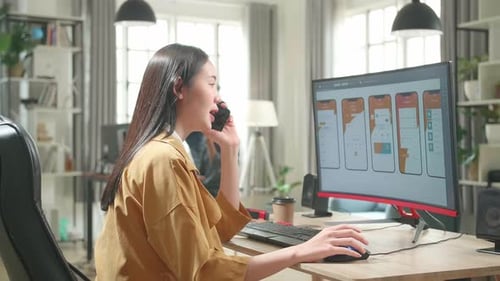 Female Mobile Application Developer Works And Talking On Mobile Phone With Graphics On Computer
