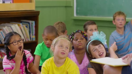 Kids listen to teacher read book and raise hands in school classroom