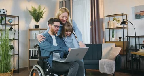 Hombre en silla de ruedas aplicando una computadora portátil junto con su hija y su esposa bastante alegre en casa