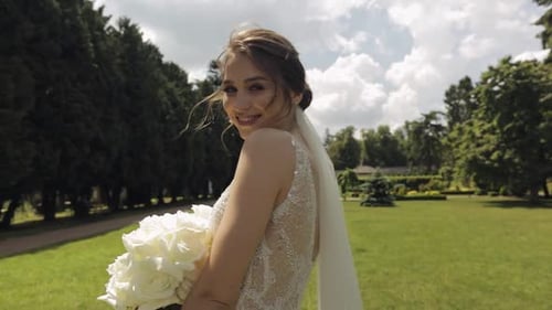 Beautiful Stylish Bride in White Wedding Dress and Veil Holding Wedding Bouquet in Hands in Park