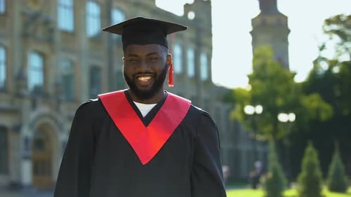 Satisfied Black Male in Graduation Gown Smiling Camera on University Background