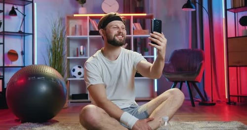 Hombre sentado en postura de loto en el suelo después del entrenamiento en casa y disfrutando de un agradable videochat