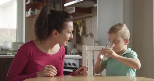 Kid Is Drinking A Glass Of Water With Mother Sitting Close By At The Kitchen