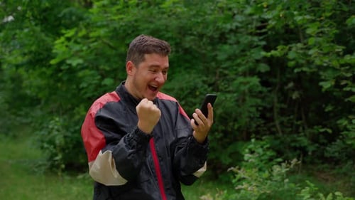 Excited happy man looking at smartphone in forest. Young adult 30s person with positive emotions