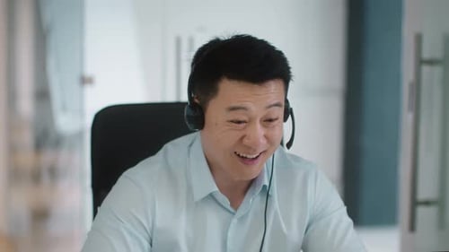 Positive Middle Aged Asian Businessman Wearing Headset with Mic Enjoying Video Conference with