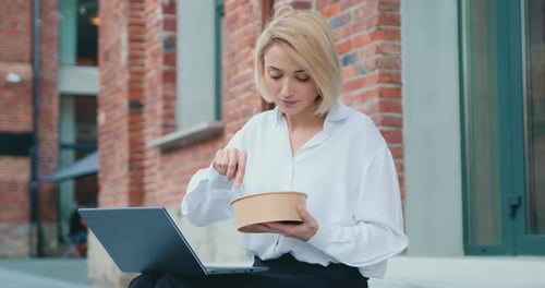 Frau beim Mittagessen mit einem Salat, während sie mit einem Laptop auf der Treppe sitzt.