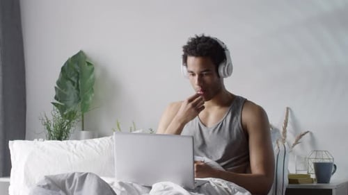 Man Works on Laptop in Bed With Headphones