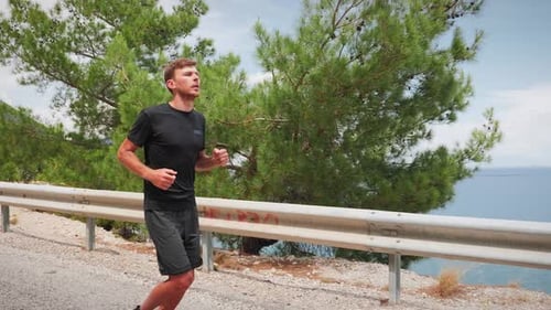 Athlete running along mountain road with sea on background.