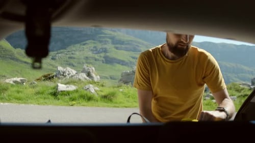 Smiling Man putting backpack in open car trunk for journey against mountain landscape