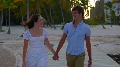 Man and woman holding hands walk on wood pier during golden hour sunset at tropical island destinati