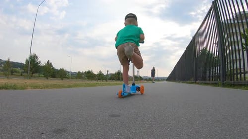 Little Boy Rides Three-wheeled Scooter and His Older Sister on Skateboard Outdoor in the Park