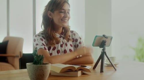 Mujer joven sentada frente al teléfono en un trípode grabando un Vídeo educativo