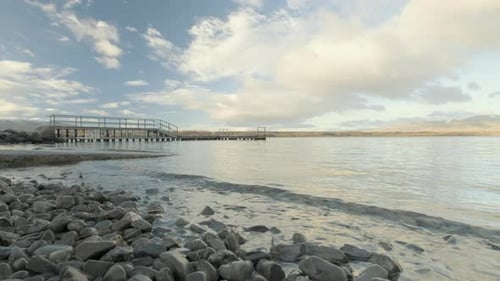 Niedrigwinkelaufnahme des Wassers aus einem See, der ins Ufer rollt, mit einem Steg im Hintergrund auf einem sonnigen