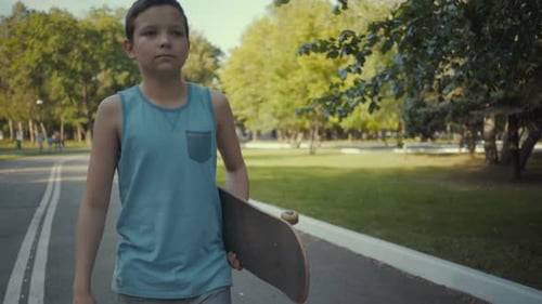 Skateboarder Walks in a Park with a Skateboard in Hands