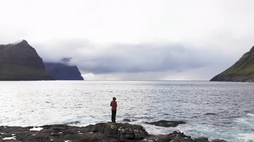 Aerial View of Unrecognizable Woman Stands on Rock Cliff Looking at Wilderness Background of Faroe