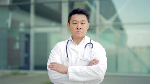 Close up portrait of Asian doctor looking at camera with arms crossed on background